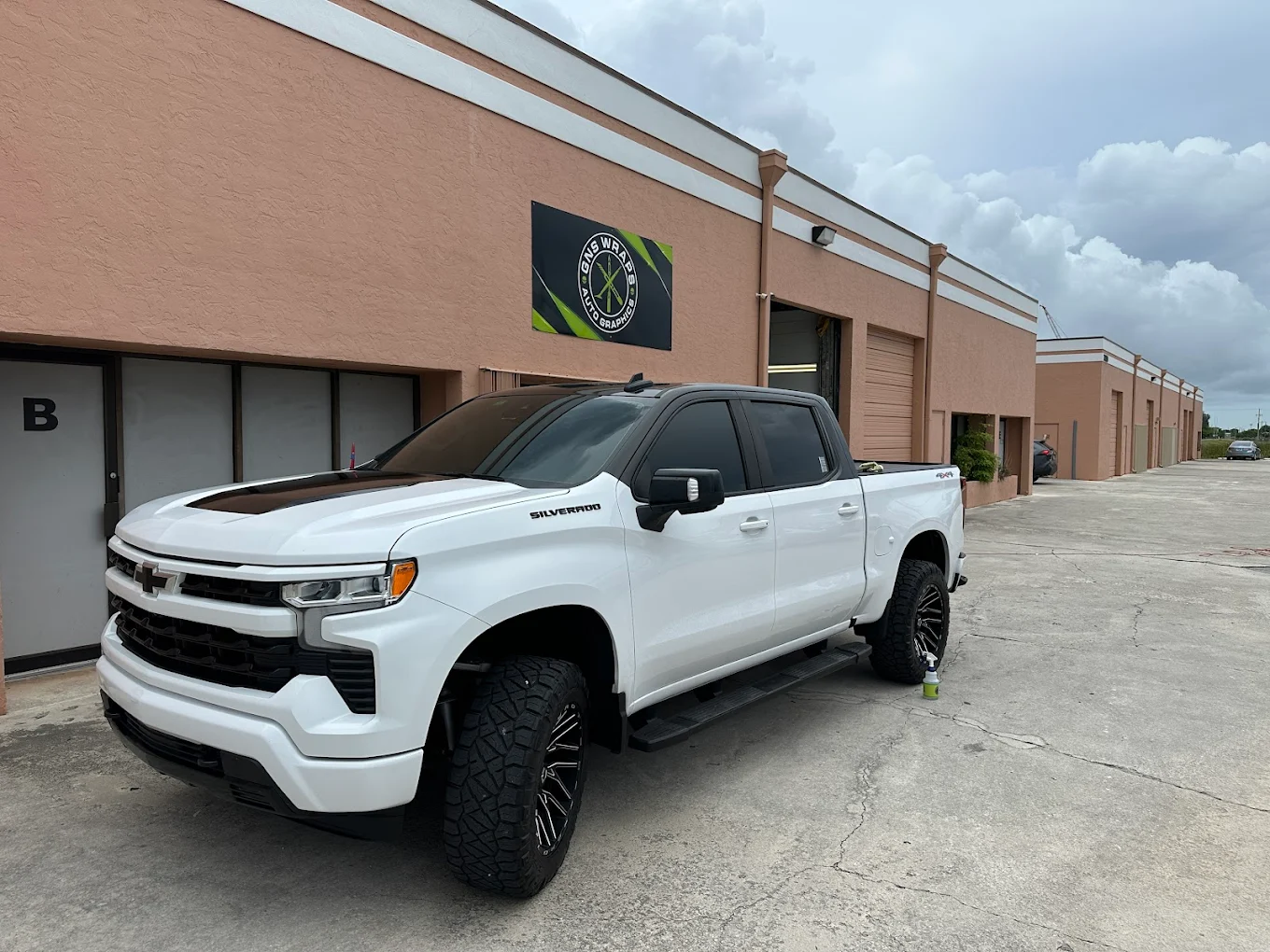 A white Chevrolet Silverado pickup truck with large black wheels is parked in front of a light brown commercial building with a sign and several garage doors. The sky is cloudy.